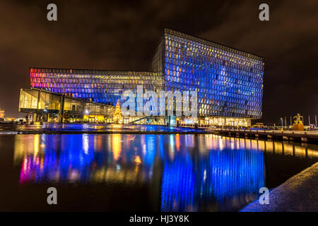 Harpa concert hall in Iceland at night lights up in multiple colors, reflecting on a pool at the front of the building. Stock Photo