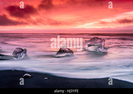 An iceberg along the shore of Jokulsarlon glacial lagoon during a vibrant red sunrise rests motionless as it is framed by cold ocean water. Stock Photo