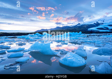 Icebergs along the shore of Jokulsarlon glacial lagoon during a blue overcast day rest motionless while framed by cold ocean water. Stock Photo