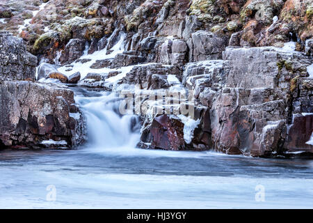 A waterfall surrounded by ice and snow in the highlands of Iceland framed rugged terrain offers scenic landscape epitomizing the frozen wilderness. Stock Photo