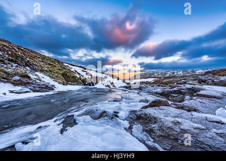 A cold snowy river in the highlands of Iceland framed by pastel skies and rugged terrain offers scenic landscape epitomizing the frozen wilderness. Stock Photo