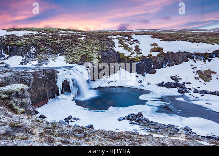 A cold snowy waterfall in the highlands of Iceland framed by pastel skies and rugged terrain offers scenic landscape epitomizing the frozen wilderness Stock Photo
