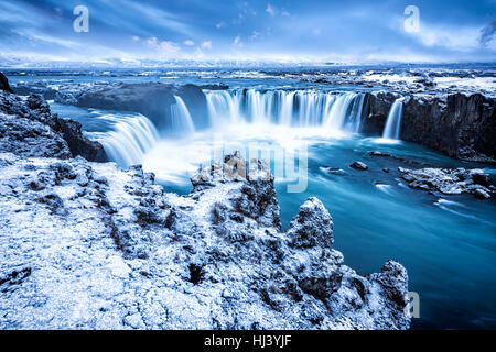 Godafoss Falls during Sunrise shows the water pouring over the edge and kicking up a misty cloud over the water with snow covering the cliffs. Stock Photo
