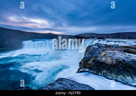 Godafoss Falls during Sunrise shows the water pouring over the edge and kicking up a misty cloud over the water with snow covering the cliffs. Stock Photo