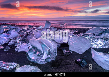 An iceberg along the shore of Jokulsarlon glacial lagoon during a vibrant red sunrise rests motionless as it is framed by cold ocean water. Stock Photo