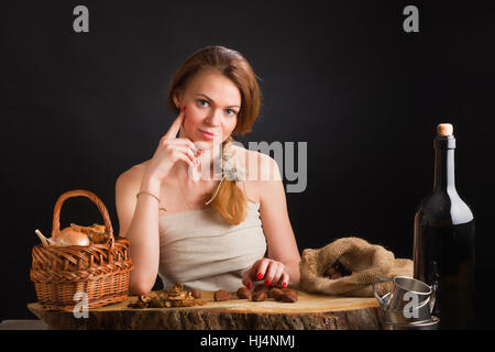 The young beautiful girl in a sundress from  canvas sits at an oak table about  basket with onions and garlic, dried mushrooms  chestnuts Stock Photo