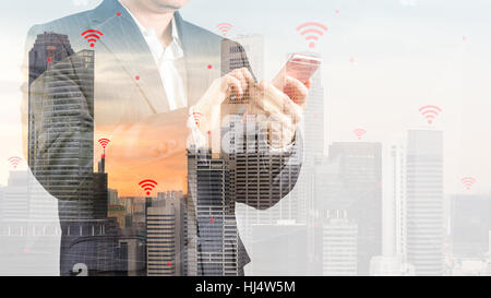 Concept of wireless connection and internet of things, IOT, shown by businessman in suit using smartphone and city as background with double exposure. Stock Photo