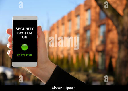 Woman holding a smartphone with home security system panel on blurred house background Stock Photo