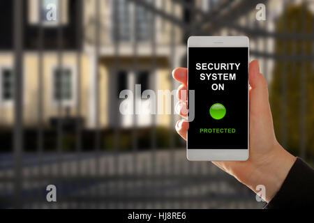 Woman holding a smartphone with home security system panel on blurred house background Stock Photo