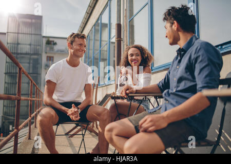 Three young friends together at outdoor cafe. Multiracial group of young people sitting around a small cafe table chatting and smiling. Stock Photo