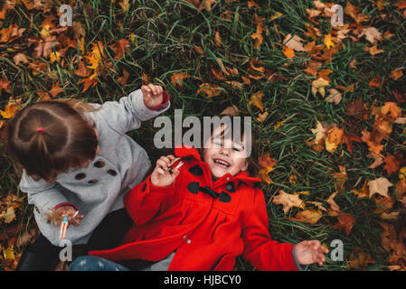 Overhead portrait of girl and toddler sister lying on grass and autumn leaves Stock Photo