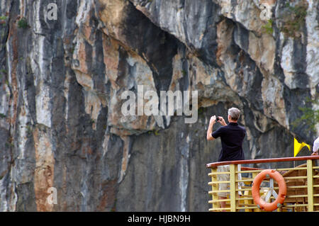 Western Tourist takes iPhone Photo of HaLong Bay, Vietnam while Standing on the Top Deck of Boat with a Rock Face Island in Backdrop Stock Photo