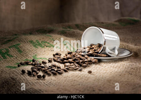 A studio, still life of a cup of coffee on a saucer with a cinnamon stick, roasted beans and biscuits on a hessian mat Stock Photo