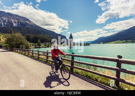 Senior woman cycling by lake with Curon bell tower, Vinschgau Valley, South Tyrol, Italy Stock Photo