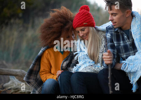 Three adult friends sitting huddled together on beach at dusk Stock Photo