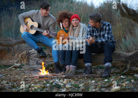 Four young adult friends playing guitar by campfire on beach at dusk
