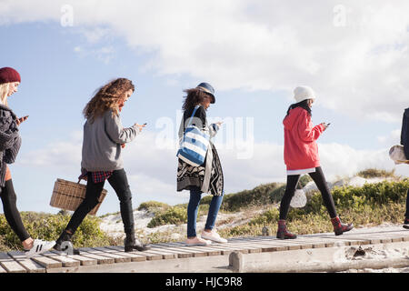 Row of young adult friends strolling along beach boardwalk reading smartphones, Western Cape, South Africa Stock Photo