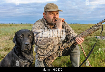 A wildfowler, or duck hunter, with his dog on the Lincolnshire marsh blowing into a duck call Stock Photo