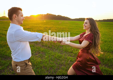 young boy in white shirt and girl in red dress whirls in field at sunset. couple spinning on the grass Stock Photo