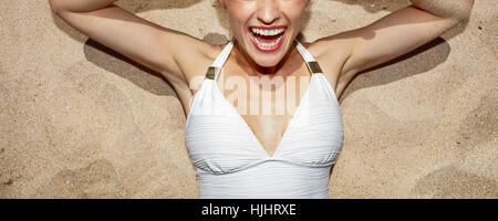 Warm sand treatment. Closeup on cheerful woman in swimsuit with funny pineapple glasses laying on the sand Stock Photo