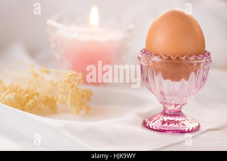 Brown fresh egg in crystal pink cup,burning candle,spring delicate yellow flowers on white linen cloth background,table setting Stock Photo
