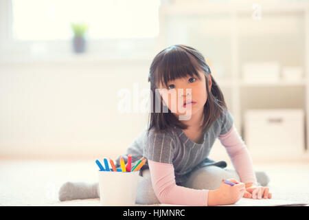 Pretty happy little japanese girl lying and drawing with pencils on white capet at home Stock Photo