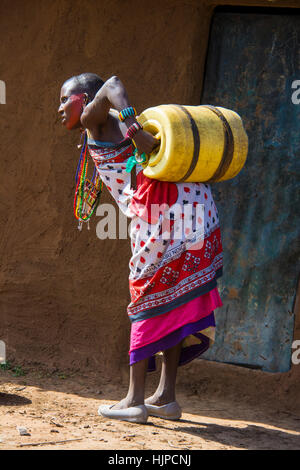 Maasai Woman struggling with a water barrel on back, wearing traditional attire, in a village near the Masai Mara National Park, Kenya, East Africa Stock Photo