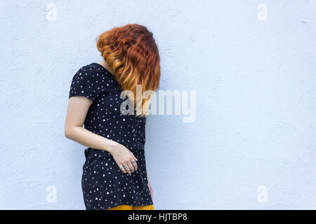 Young caucasian female with short red hair wearing black dress stands against grey wall with face covered Stock Photo