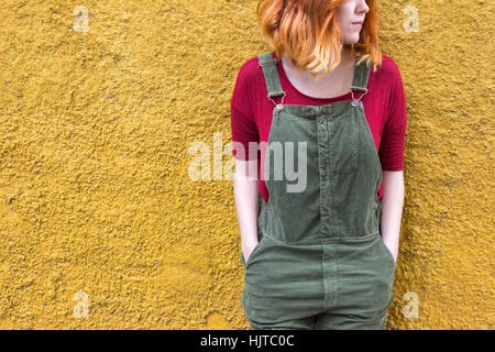 Young female with red short hair and wearing green overalls with red shirt smiles while standing against yellow Stock Photo