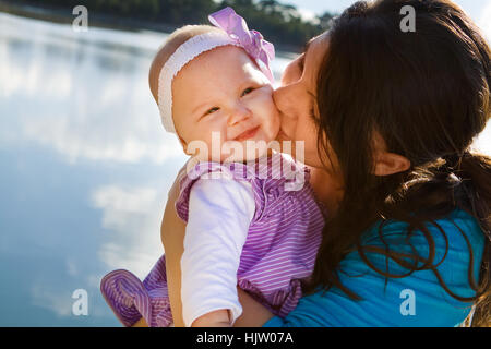 Cute smiling baby receiving a kiss from her mom by a lake Stock Photo