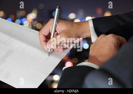 Businessman's Hand holding a pen signing to the document on blurred abstract night bokeh background as Approval Concept. Stock Photo