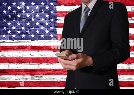 Business man hand touch screen smart phone and brick Walls are painted United States flag (USA) as communication with mighty nation concept. Stock Photo