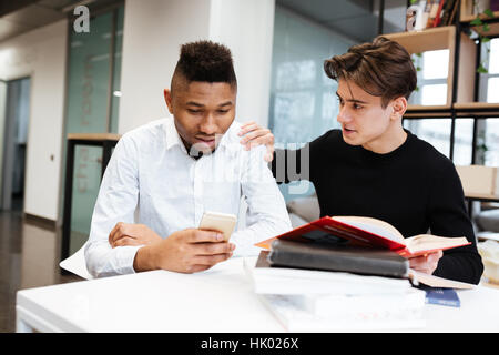 Picture of two young students sitting in library and choose between the book and cellphone. Stock Photo