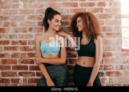 Portrait of two fitness females in gym relaxing and talking after workout. Young woman friends standing against wall and smiling. Stock Photo