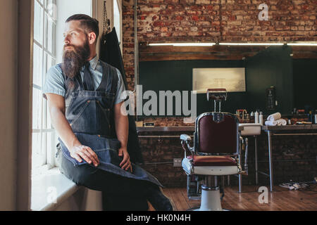 Young barber at his barbershop. Caucasian male with long beard sitting on sill of a window and looking outside. Stock Photo