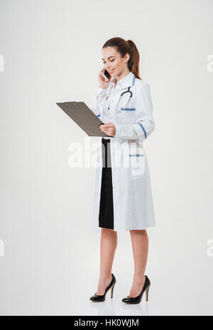 Full length of happy young woman doctor holding clipboard and talking on mobile phone over white background Stock Photo