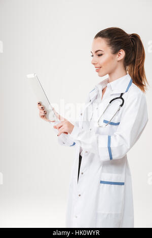 Cheerful lovely young woman doctor smiling and using tablet over white background Stock Photo