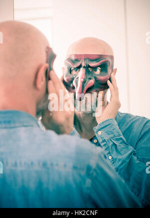 Old man cover face with carnival mask. Looking at himself in a mirror. Concept of ageing, self image as a senior person and hiding identity. Stock Photo