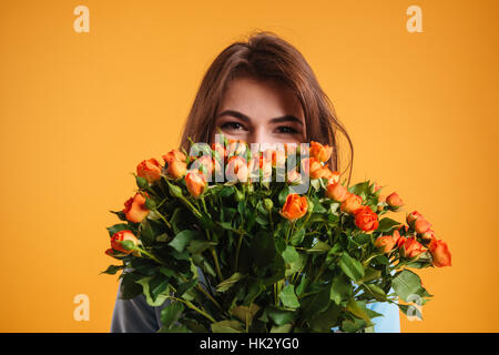 Smiling cute young woman standing and hiding behind bouquet of flowers over yellow background Stock Photo