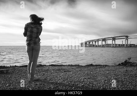 Girl stands on a sea coast, modern bridge under construction on a horizon, black and white photo Stock Photo