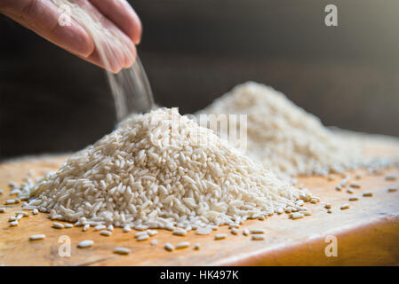 Hand pouring rice on pile of white rice on wooden table background, metaphor ingredient, nutrition, carbohydrate food concept, selective focus Stock Photo