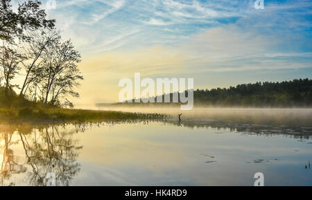 Brilliant and bright mid-summer sunrise on a lake.   Warm water and cooler air at daybreak creates misty fog patches.  Still water on calm waters. Stock Photo