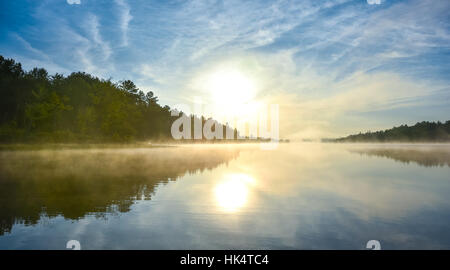 Brilliant and bright mid-summer sunrise on a lake.   Warm water and cooler air at daybreak creates misty fog patches.  Still water on calm waters. Stock Photo