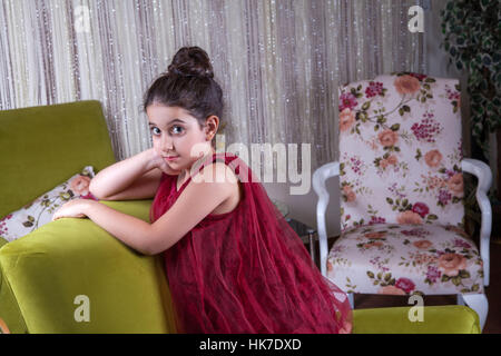 Cute lovlely middle eastern girl with dark red dress and collected hair posing and liying on green sofa at home interior. Stock Photo