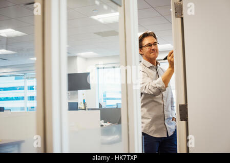 A man standing in an office writing on a whiteboard, seen through a doorway. Stock Photo