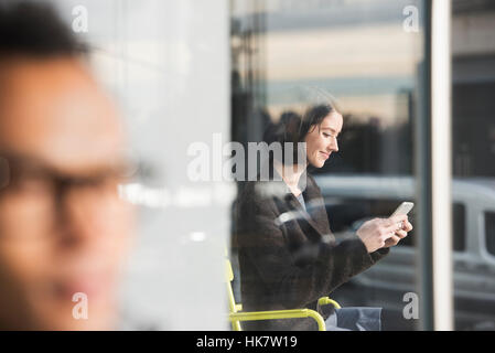 A seated woman seen through a window looking at a cellphone, with an out-of-focus man in the foreground. Stock Photo