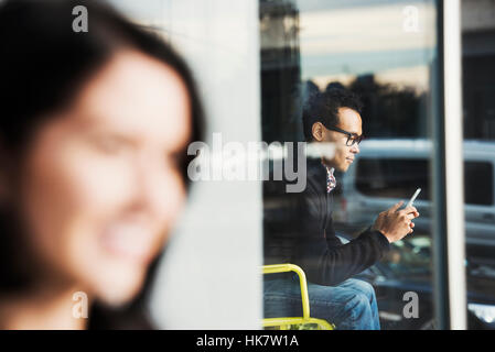 A seated man seen through a window looking at a cellphone, with an out-of-focus woman in the foreground. Stock Photo