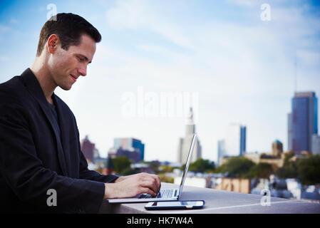 A young man standing on a rooftop working at an open laptop with a cellphone beside it. Stock Photo