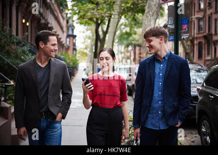 Two young men and a young woman walking down a city street looking at a cellphone. Stock Photo