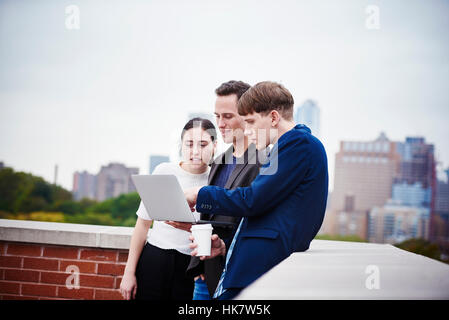 A young woman and two young men standing on a rooftop looking down at a laptop computer together. Stock Photo